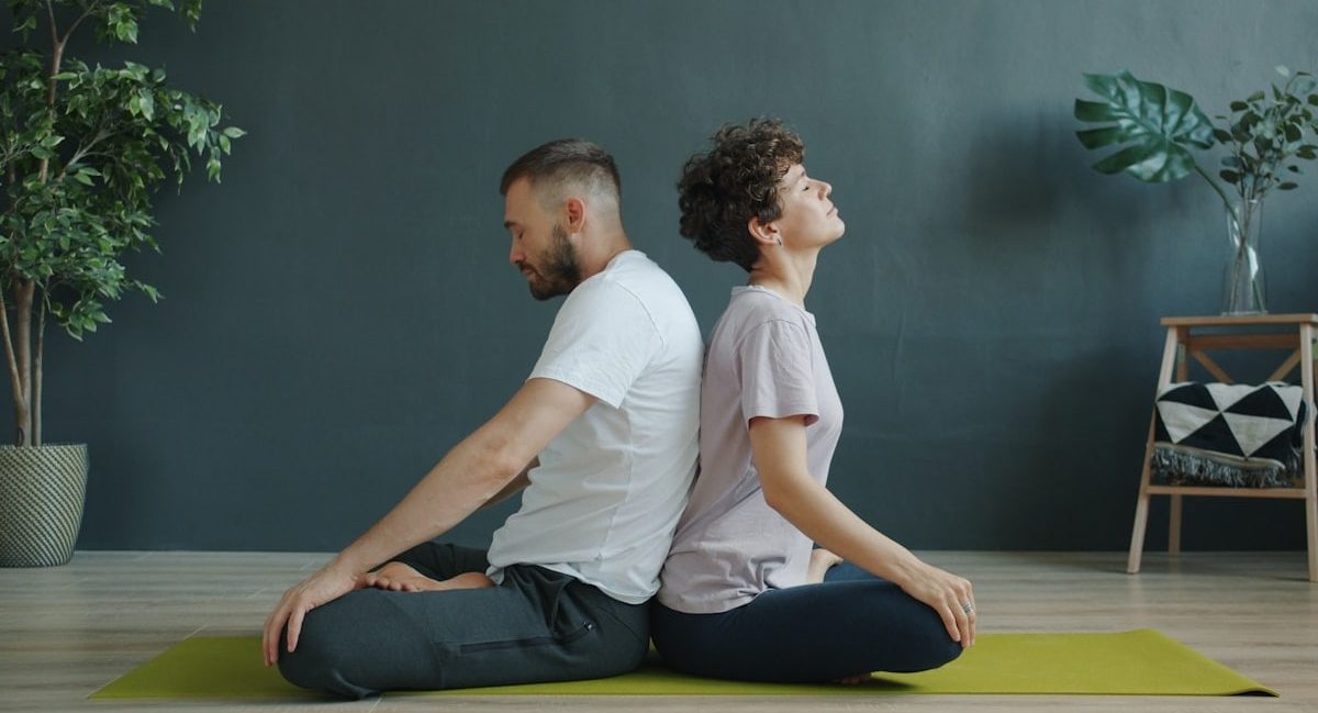 Man and woman are practising pair yoga doing warm-up exercises sitting back to back on mat in studio. Healthy lifestyle, bodycare and pairwork concept.